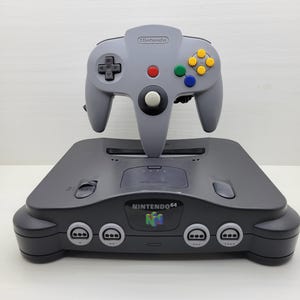 May include: A charcoal grey Nintendo 64 video game console with a front-facing cartridge slot and four controller ports. A light grey wired controller with a central analogue stick, a red start button, and yellow, green, and blue buttons sits on top.