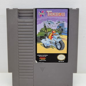 Peut inclure: Une cartouche de jeu grise pour Nintendo Entertainment System du jeu Thundercade par Sammy. L'étiquette présente une illustration colorée d'une moto, d'un char, d'un sous-marin et d'un jet. Elle inclut le texte Licensed by Nintendo et Made in Japan.