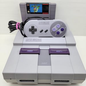 May include: A grey Super Nintendo console with a game cartridge labelled "M. ROWORLD" and a matching controller. The console has purple buttons, and the controller has a directional pad and purple buttons. The game cartridge is inserted into the console.