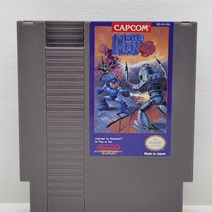 May include: A grey Nintendo Entertainment System cartridge for Mega Man 3. The label shows a colourful illustration of the blue Mega Man character fighting a robot. The Capcom logo is at the top, with "Made in Japan" at the bottom.