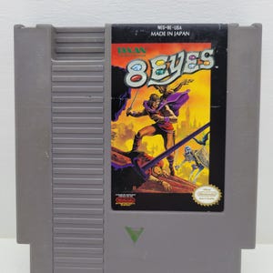 Peut inclure: Une cartouche de jeu grise pour Nintendo Entertainment System intitulée 8 Eyes. L'étiquette illustre un guerrier à cape violette avec un oiseau et un ennemi squelette. Le texte inclut Taxan, 8 Eyes, NES-8E-USA et Made in Japan.