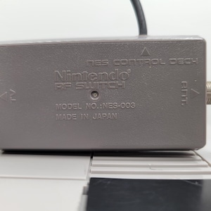 May include: A grey Nintendo RF Switch, model NES-003, with the text "NES Control Deck" and "Made in Japan". The switch has a coaxial connector and a black cable.
