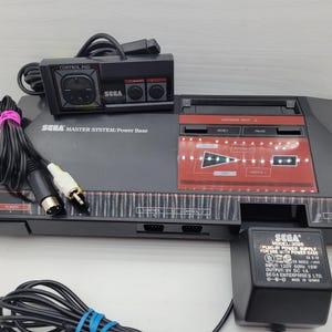 May include: A black Sega Master System video game console with a red accent panel, a black controller, and a power supply. The console has the text "SEGA MASTER SYSTEM/Power Base" printed on it. The controller is labeled "CONTROL PAD".