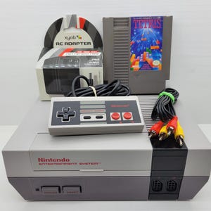 May include: A Nintendo Entertainment System console with a controller, game cartridge, and AC adapter. The console is grey and black, with red and grey buttons. The game cartridge is labelled "Tetris".