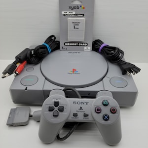 May include: A gray PlayStation console with a matching controller, cables, and a memory card. The controller has a directional pad, buttons, and the PlayStation logo. The memory card is labeled "1MB".