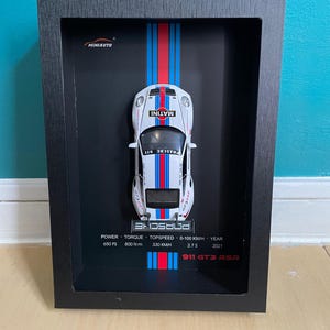 Op de afbeelding: Een ingelijste miniatuur witte Porsche 911 GT3 RSR raceauto met blauwe en rode strepen. De auto wordt tentoongesteld tegen een zwarte achtergrond met gedrukte specificaties. De lijst is zwart.