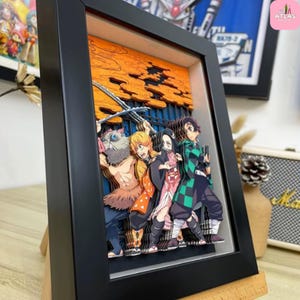 May include: A framed 3D paper art piece featuring characters from the anime series Demon Slayer. The artwork depicts several characters in vibrant colors, set against an orange and blue background. The frame is black and the piece is displayed on a wooden stand.