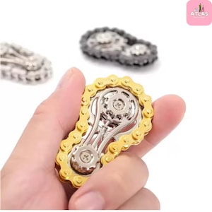 以下が含まれることがあります： 自転車のチェーンの形をした金と銀のフィジェットスピナーが手に持たれています。背景には、他の2つの同様のスピナーがあります。スピナーは金色のチェーンと銀色のギアを持っています。