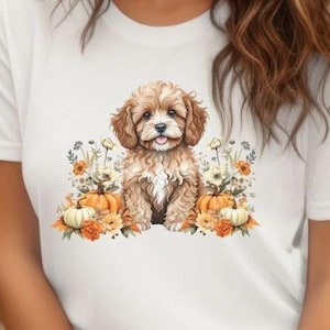 May include: White t-shirt featuring a watercolor illustration of a fluffy brown and white puppy. The puppy is surrounded by pumpkins, flowers, and foliage in shades of orange, yellow, and green, creating a fall theme.