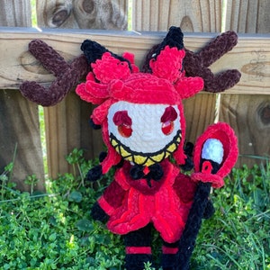 May include: A red crocheted plush toy with black horns and a black staff. The toy has a white face with red eyes and a yellow smile. The toy is sitting on a bed of green grass.