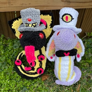 May include: Two crocheted plushies with unique designs. One is yellow and black with a red eye and a gray top hat. The other is purple and white with a yellow eye and a white top hat.