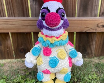 Könnte beinhalten: Eine gehäkelte Clownpuppe mit einem weißen Körper, einem roten Hut, einem violetten Gesicht und einer bunten Polka-Dot-Hose. Die Puppe hat eine rosa Nase und schwarze Augen.
