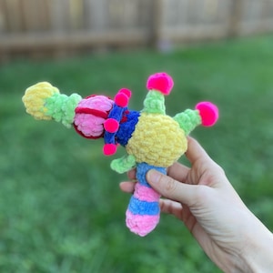 May include: A colorful, handmade crochet toy gun. The toy features a combination of bright colors, including yellow, green, pink, blue, and red. The design includes a handle with pink and blue stripes, and a barrel with a variety of shapes.