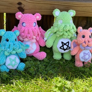 Könnte beinhalten: Vier gehäkelte Plüschtiere in Form von Cthulhu mit verschiedenen Tentakeln und Flügeln in verschiedenen Farben. Die Spielzeuge sind rosa, grün, blau und orange. Jedes Spielzeug hat einen weißen Stern mit einem schwarzen Pentagramm in der Mitte auf der Brust.