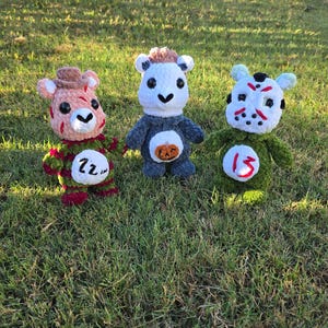 May include: Three plush crochet dolls with soft yarn textures. The left doll wears a brown hat and red and green striped sweater with the text 22. The center doll is white and gray with a pumpkin patch. The right doll wears a white hockey mask with red markings and the number 13 on its green body.