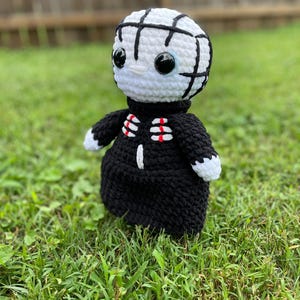 Puede incluir: Muñeca de ganchillo hecha a mano que se asemeja a un personaje de una película de terror. La muñeca tiene una cabeza blanca con líneas negras, ojos negros grandes y un atuendo negro y rojo. La muñeca está sobre hierba verde.