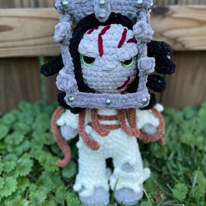 May include: A crocheted plush toy of a character wearing a grey cage with metal accents. The character has grey skin, red stripes, and black hair. The toy is standing on green grass.