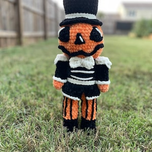 May include: A handmade crochet pumpkin doll wearing a black top hat and a black and white outfit. The pumpkin has a smiling face with black eyes and a black mouth. The doll is standing on green grass.