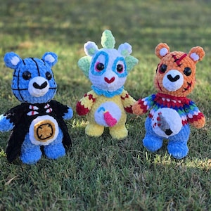 May include: Three plush crochet bears with distinct designs. The left bear is blue with black grid lines and a black cape. The center bear is yellow with green accents and a pink drink icon. The right bear is orange with a red sweater and knife icon.