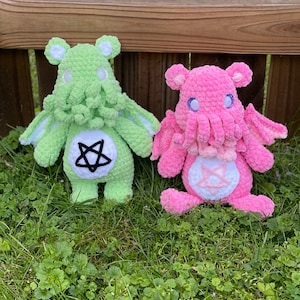 Puede incluir: Dos peluches de crochet con forma de osos con tentáculos. El oso de la izquierda es verde con un pentagrama blanco en el pecho. El oso de la derecha es rosa con una estrella blanca en el pecho.