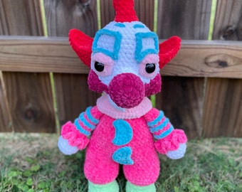 Könnte beinhalten: Eine gehäkelte Clownpuppe mit einem rosa-weißen Outfit, roten Haaren und blauen Augen. Die Puppe trägt ein blau-weiß gestreiftes Hemd und hat eine rosa Nase.