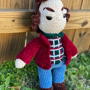 Può includere: Giocattolo di peluche all'uncinetto di un personaggio che indossa una giacca rossa, una camicia a quadri verde e bianca, jeans blu e scarpe marroni. Il giocattolo ha i capelli castani e un'espressione seria.