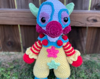 Könnte beinhalten: Eine gehäkelte Clownpuppe mit grünem Hut, roter Nase und blau-gelb gestreiftem Outfit. Die Puppe hat große schwarze Augen und ein rosa-violettes Gesicht.