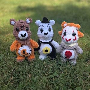 Puede incluir: Tres peluches de ganchillo hechos a mano colocados uno al lado del otro. El juguete de la izquierda es un oso marrón y naranja con detalles cosidos. El del medio es un oso blanco y negro con un pequeño sombrero negro. El de la derecha es un oso gris y naranja.