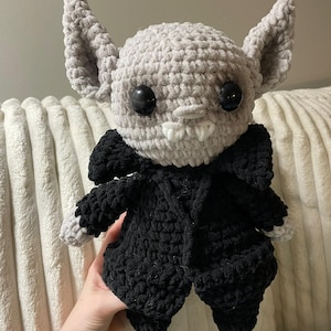 Puede incluir: Muñeca de murciélago de ganchillo hecha a mano con cabeza y orejas grises, ojos negros grandes y colmillos blancos. El murciélago lleva un atuendo negro. La muñeca es suave y afelpada, ideal como juguete o artículo decorativo.