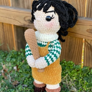 May include: A crocheted doll with black hair, wearing a green and white checkered shirt, a yellow overall dress, and brown boots. The doll is holding a wooden baseball bat.