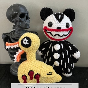 May include: Three crocheted plush toys: a black and white bear with a red smile, a yellow duck with red dripping paint, and a black skeleton. The toys are on a black surface with the text "PDF ONLY" in white.