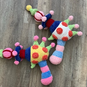 Puede incluir: Tres juguetes de sonajero de crochet con forma de corona con un mango a rayas rosa, azul y blanco. Las coronas están decoradas con lunares rojos, amarillos y verdes.