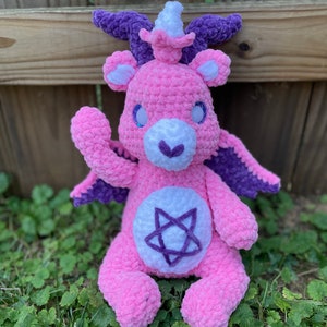 May include: A pink crocheted Care Bear with purple wings, a horn, and a pentagram on its chest. The bear is sitting on a bed of green grass.