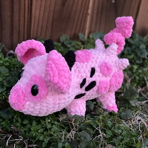 May include: A pink and black crocheted pig plush toy with black eyes. The pig is sitting on a bed of green grass.