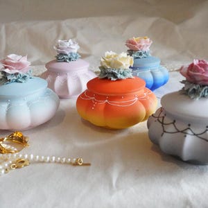 May include: Five ceramic trinket boxes with floral accents. Each box is a different colour, including pink, blue, orange, white, and light blue. The boxes are decorated with delicate floral designs and have a vintage aesthetic.
