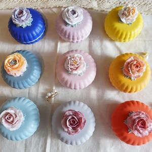May include: Ten small round ceramic boxes with floral designs. Each box has a different colour and floral pattern. The boxes are arranged in a circle on a white surface.