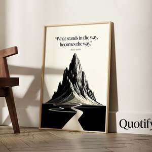 May include: A framed black and white illustration of a mountain with a winding road leading to the peak. The quote "What stands in the way, becomes the way." by Marcus Aurelius is printed below the image. The word "Quotify" is printed in black at the bottom right corner of the image.