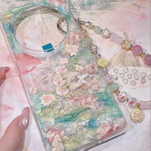 Op de afbeelding: Transparante telefoonhoes met een bloemen- en vlinderontwerp. De hoes heeft roze, witte en gouden accenten, met een kralenband in roze en groene tinten. De hoes heeft een ring om de band aan te bevestigen.