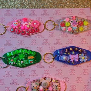 May include: A collection of personalized keychains with various designs. Each keychain features a name spelled out with colorful letters and adorned with decorative elements like flowers, bows, and cartoon characters. The keychains are on a pink background.