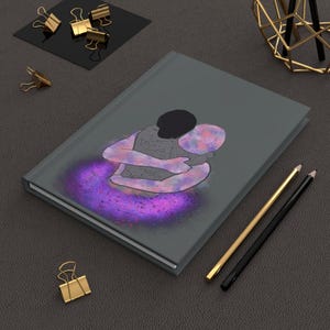 Op de afbeelding: Een grijze journal met een tekening van twee figuren die elkaar omarmen, omgeven door een paarse en roze aura. Gouden en zwarte potloden, samen met gouden paperclips, zijn verspreid rond de journal op een donkergrijze ondergrond.