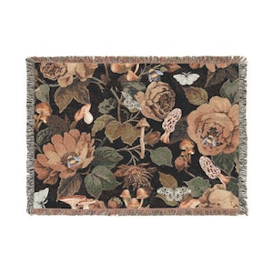 Pode incluir: Uma manta tecida com um fundo escuro com um design floral e de cogumelos. A manta tem uma borda com franjas e apresenta grandes rosas castanhas, vários cogumelos, borboletas e folhagens verdes. A paleta de cores inclui tons de castanho, castanho e verde.