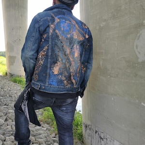 Può includere: Giacca di jeans blu con un design astratto blu e color rame sul retro. La giacca è abbinata a jeans scuri, un cappello nero e scarpe da ginnastica bianche. Una bandana nera pende dalla tasca posteriore.