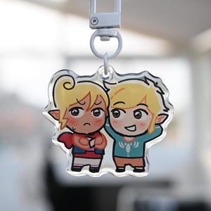 Puede incluir: Un llavero de acrílico transparente con dos personajes de dibujos animados con pelo rubio y orejas puntiagudas. Un personaje frunce el ceño con los brazos cruzados, con una capa roja y una camisa azul. El otro personaje sonríe con los brazos levantados, con una camisa azul. El llavero tiene un cierre plateado.