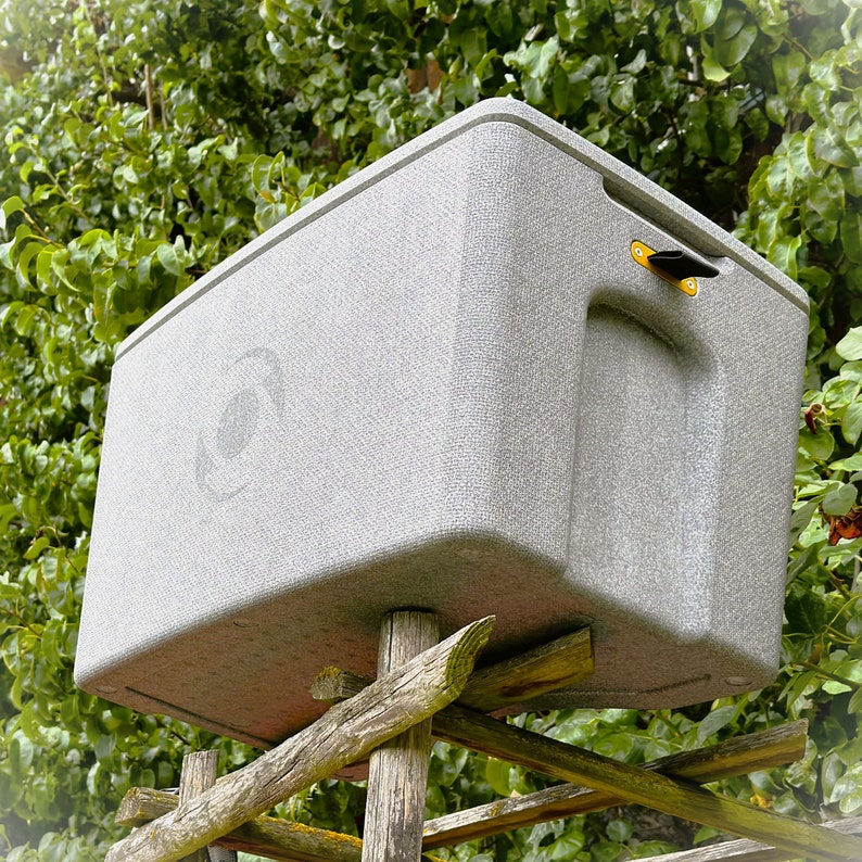 Grey cool box by cagoon, positioned on a vintage rack at an ol farm. Plants in the background. It looks like the cool box is extremely light.