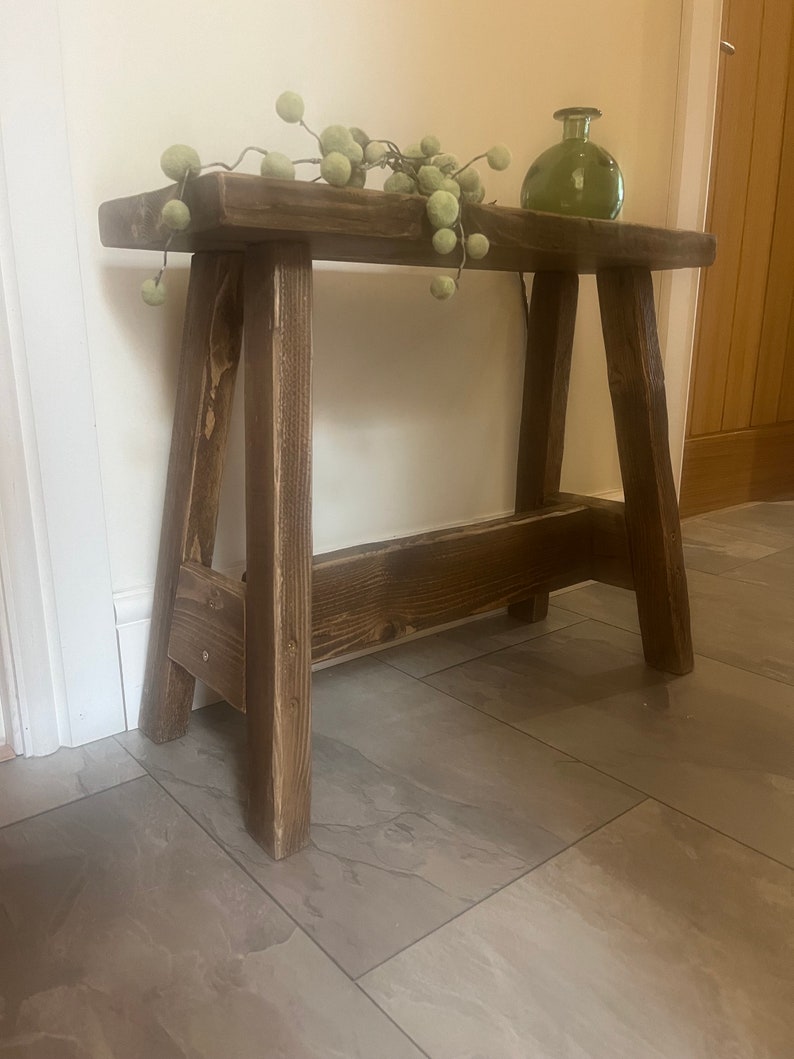 Op de afbeelding: Een rustieke houten kruk met een donkerbruine afwerking. De kruk heeft een eenvoudig ontwerp met twee schuine poten en een plat bovenblad. Een takje groene kunstgroen met ronde ballen ligt op de kruk.