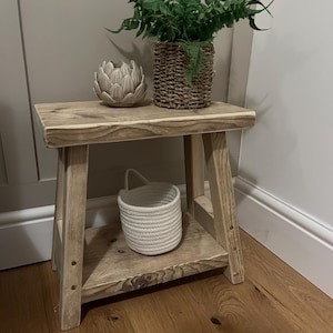 May include: A rustic wooden stool with a shelf, featuring a natural wood finish. The stool has a simple design with a rectangular top and two angled legs. A small white basket and a decorative ceramic artichoke are on the top shelf. A potted plant with green leaves is on the top of the stool.