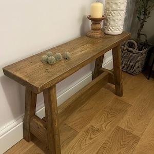May include: A rectangular wooden bench with a medium brown finish, featuring four angled legs and a horizontal support stretcher. The bench surface displays a visible wood grain texture and four small, round, green decorative spheres.