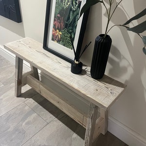 Op de afbeelding: Een witgeschilderde houten bank met een rustieke, verweerde afwerking. De bank heeft een eenvoudig ontwerp met twee poten aan elke kant en een platte bovenkant. Hij staat in een gang met een zwart ingelijst drukwerk van botanische illustraties en een zwarte vaas met een plant.