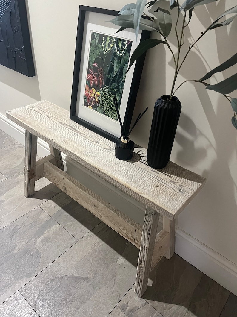 Op de afbeelding: Een witgeschilderde houten bank met een rustieke, verweerde afwerking. De bank heeft een eenvoudig ontwerp met twee poten aan elke kant en een platte bovenkant. Hij staat in een gang met een zwart ingelijst drukwerk van botanische illustraties en een zwarte vaas met een plant.