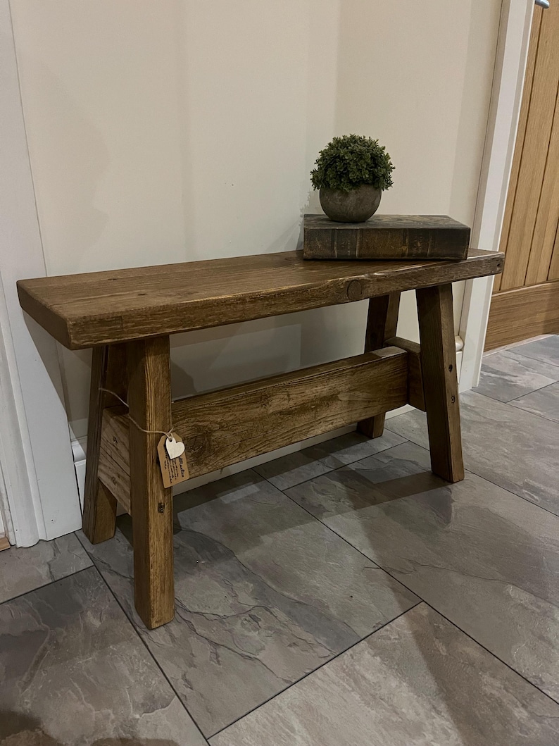 Op de afbeelding: Een rustieke houten bank met een donkerbruine vlek. De bank heeft een eenvoudig ontwerp met twee schuine poten en een enkele plank voor de zitting. De bank staat op een betegelde vloer.